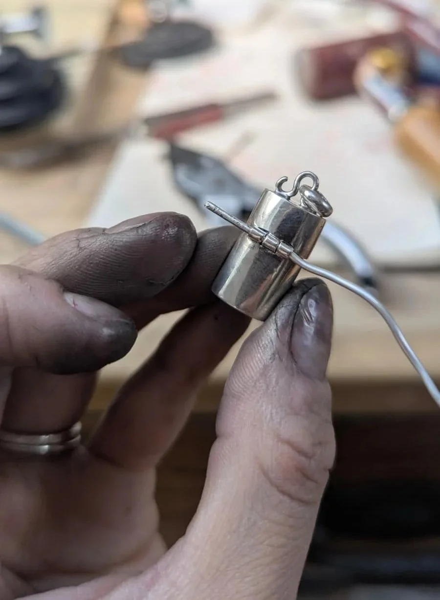 Close-up of a pair of fingers holding a small metal cylindrical object with a wire loop on top, possibly jewelry or a jewelry part, on a wooden workbench with tools and supplies blurred in the background.