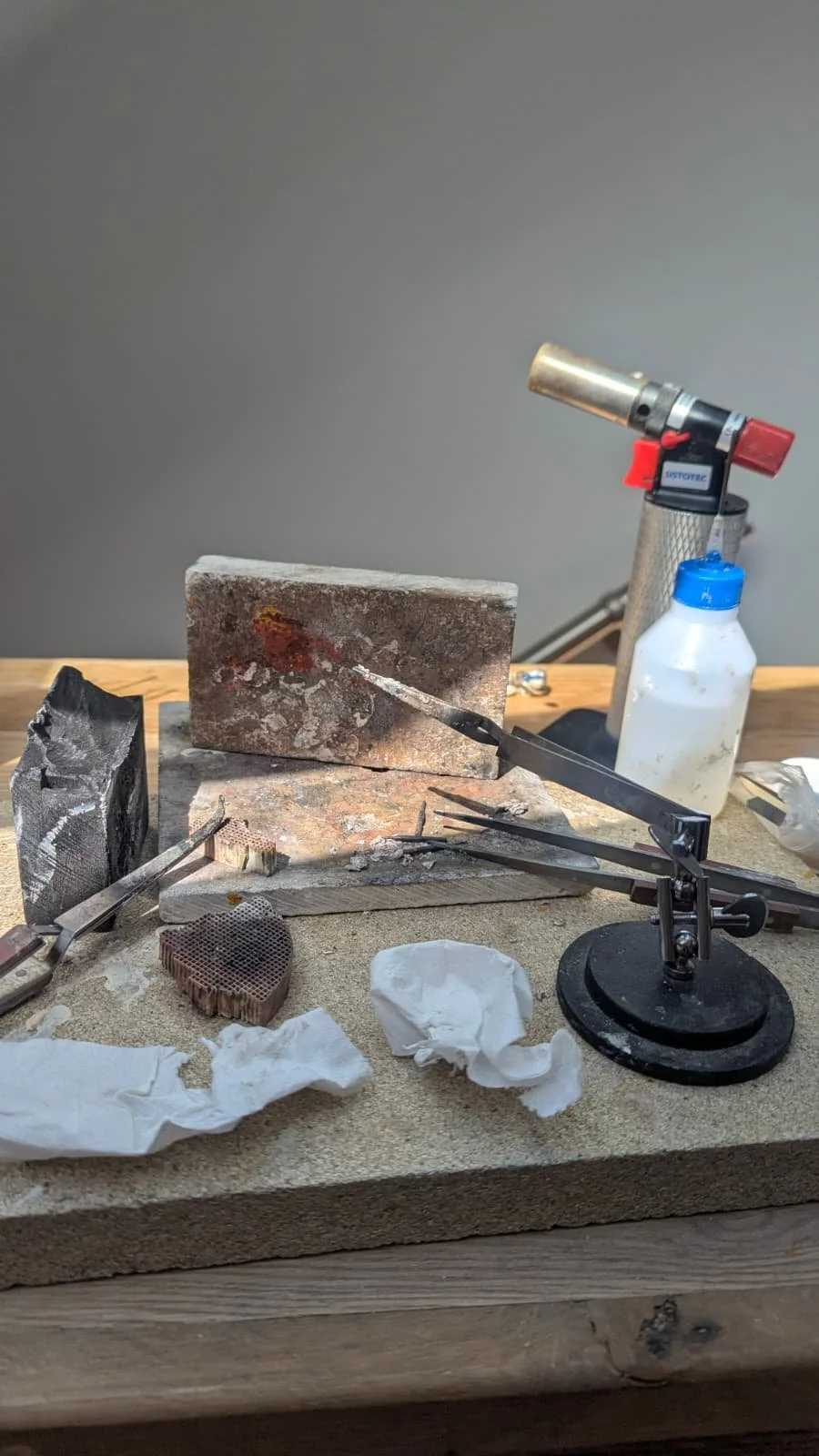 Jeweler's workbench with a glass crucible, small tools, a blowtorch, a bottle of cleaning solution, and scattered wipes and debris.