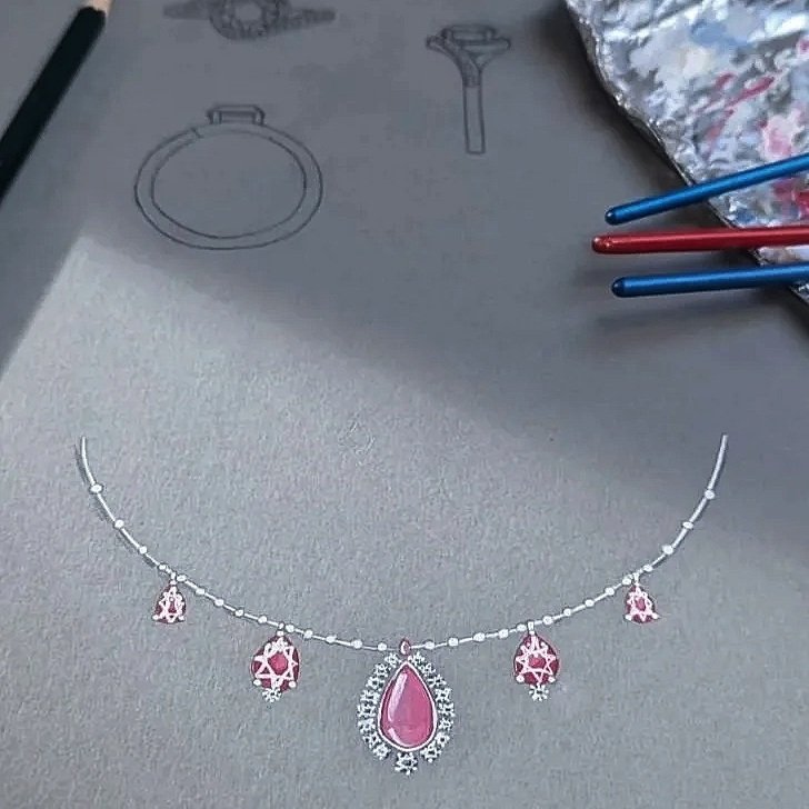 A necklace with pink gemstone pendant and matching smaller charms, laid on a gray surface with jewelry sketches and coloring tools nearby.