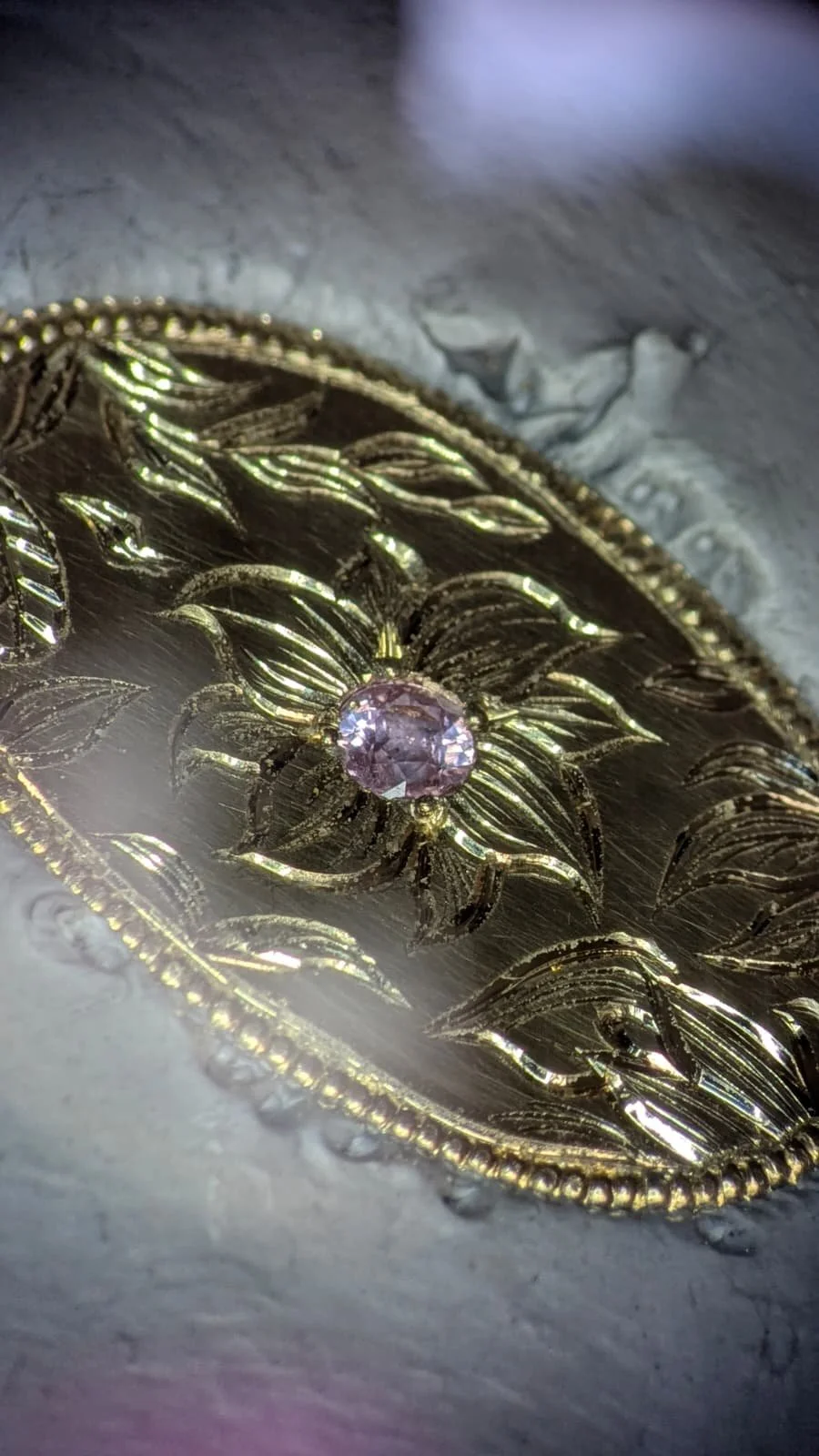 Close-up of a gold decorative brooch or jewelry piece with a purple gemstone at its center, set against a textured gray background.