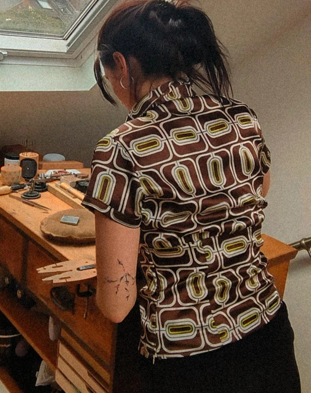 A person with dark hair in a ponytail is standing with their back to the camera, wearing a retro patterned shirt. They are leaning over a wooden desk that has various small items on it, including a small sculpture, a phone, and some tools. A tattoo of a branch with leaves is visible on their upper arm.