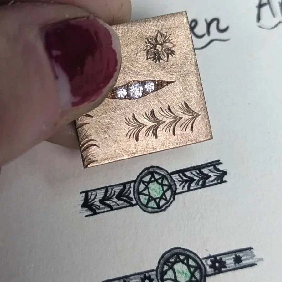 Close-up of a person's finger with dark red nail polish holding a small square stamping plate with floral and leaf designs, used for nail art. The background shows hand-drawn jewelry sketches with circular and star-shaped motifs.