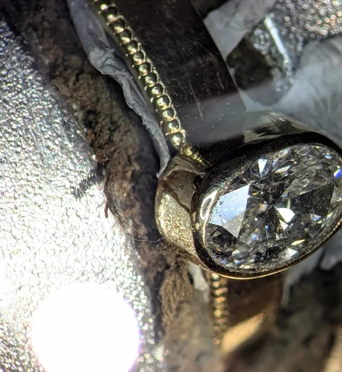 Close-up of a diamond and a gold chain set in a metallic jewelry piece with soil and dirty fabric nearby.