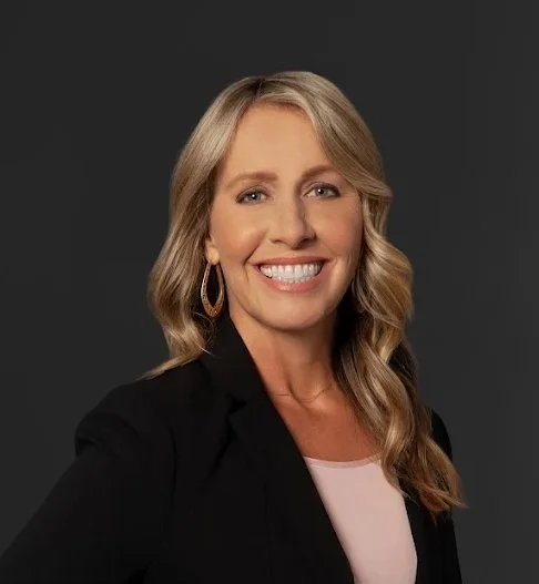 A woman with blonde hair smiling, wearing a black blazer and gold hoop earrings, against a dark background.