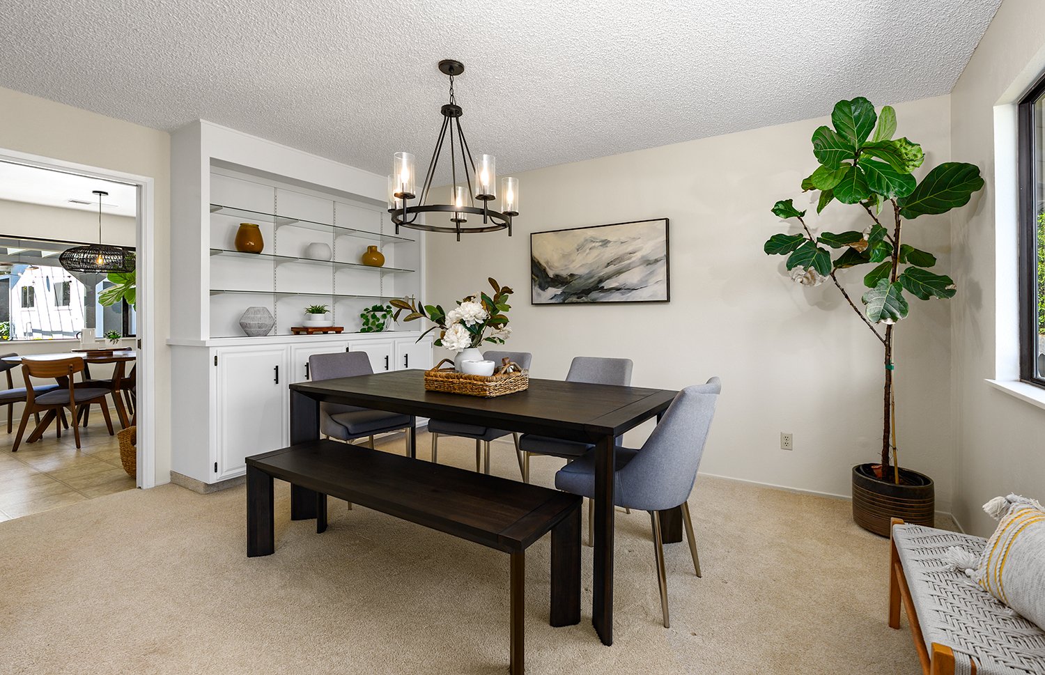 Dining room with a dark wood table, four gray upholstered chairs, a chandelier, a built-in white shelf with decor, a large potted fiddle leaf fig, and an abstract wall painting.