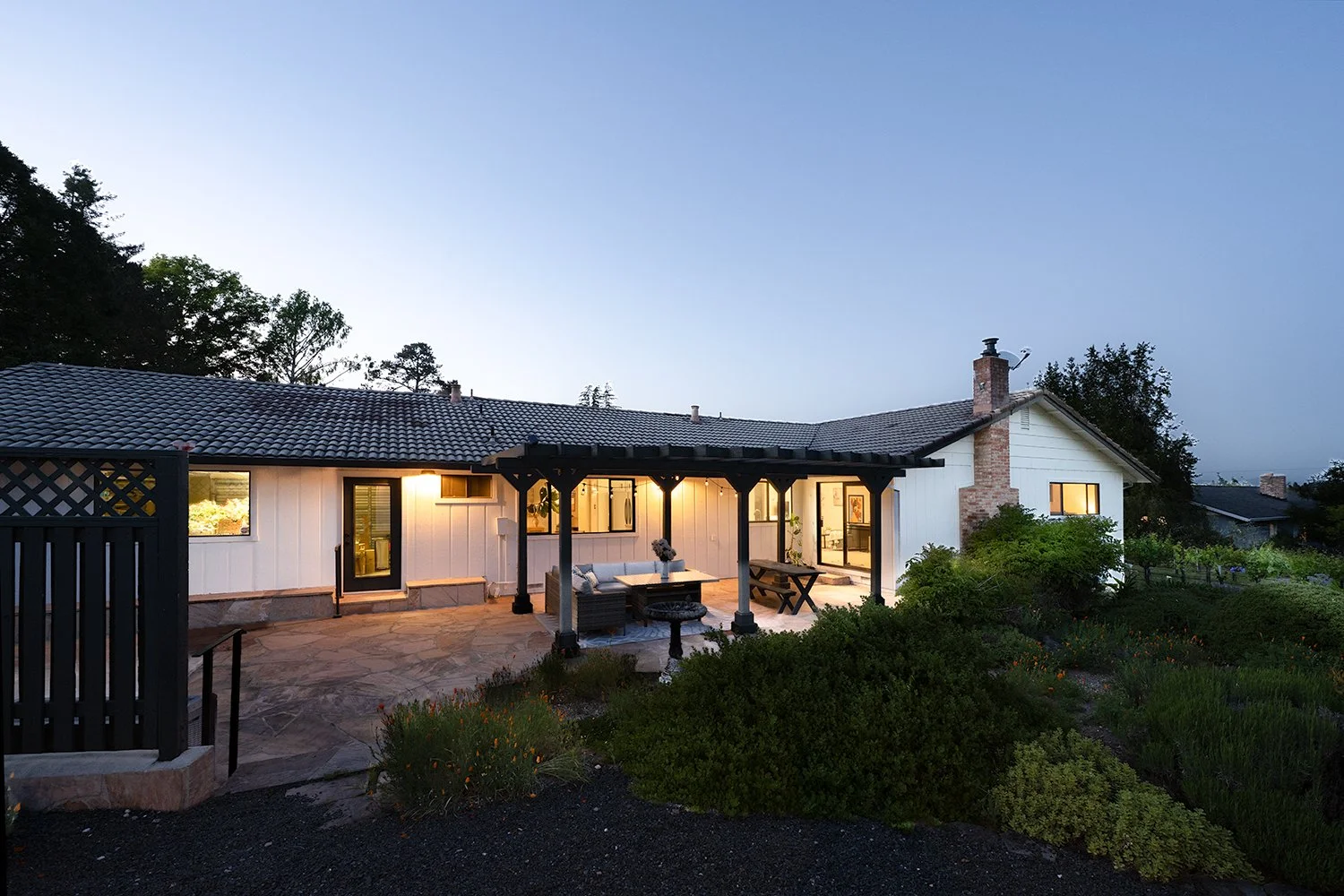 A backyard view of a white house at dusk with outdoor seating under a wooden pergola, garden plants, and illuminated windows.