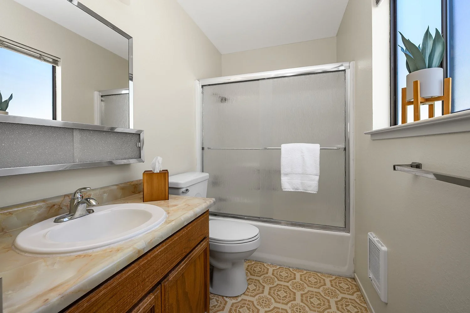 A small bathroom with a sink, mirror, toilet, shower with frosted glass doors, and a window with a plant in a white pot.