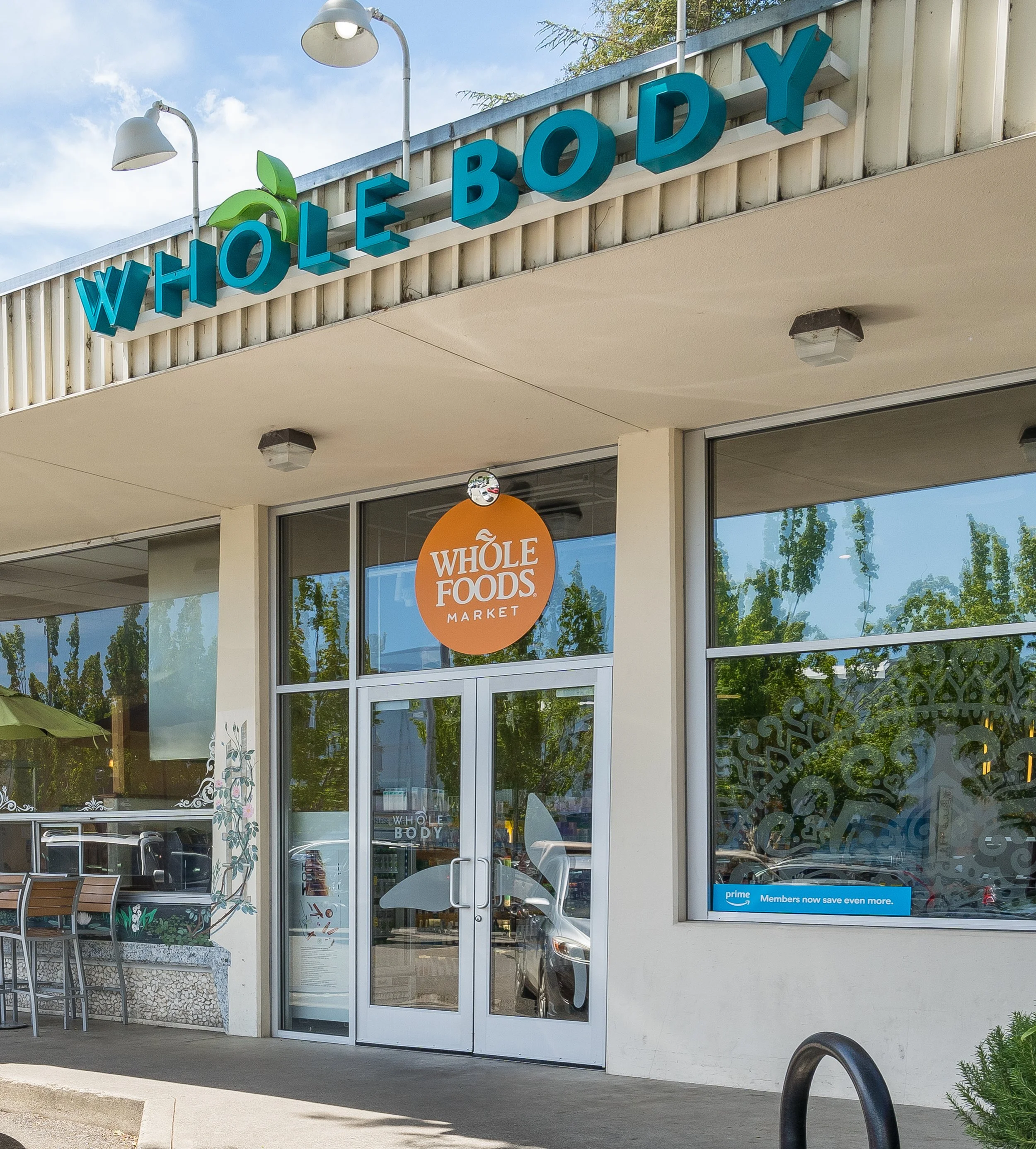Exterior view of a Whole Foods Market store entrance with blue 3D letters spelling 'WHOLE BODY' and a logo featuring green leaves. The glass doors reflect trees and parked cars outside. There is a patio with chairs and a table on the left side of the