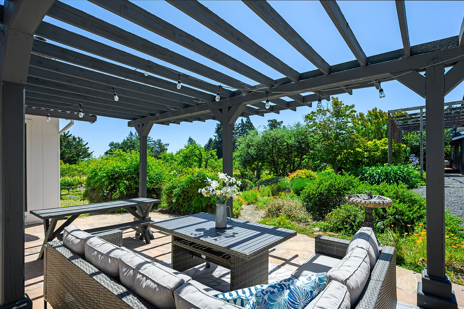 Outdoor patio area with wicker sofa, table with flowers, pergola with string lights, and garden with lush green bushes and trees under a clear blue sky.