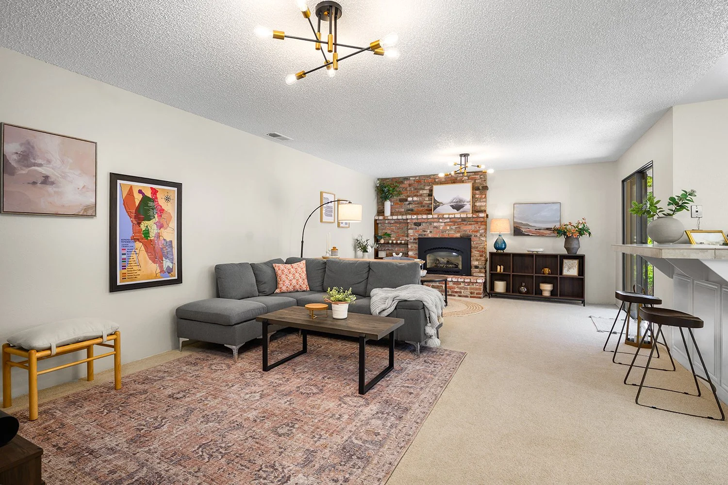 Living room with gray sectional sofa, wooden coffee table, brick fireplace, artwork on walls, modern chandelier, and barstools near a breakfast bar, decorated with potted plants and lamps.