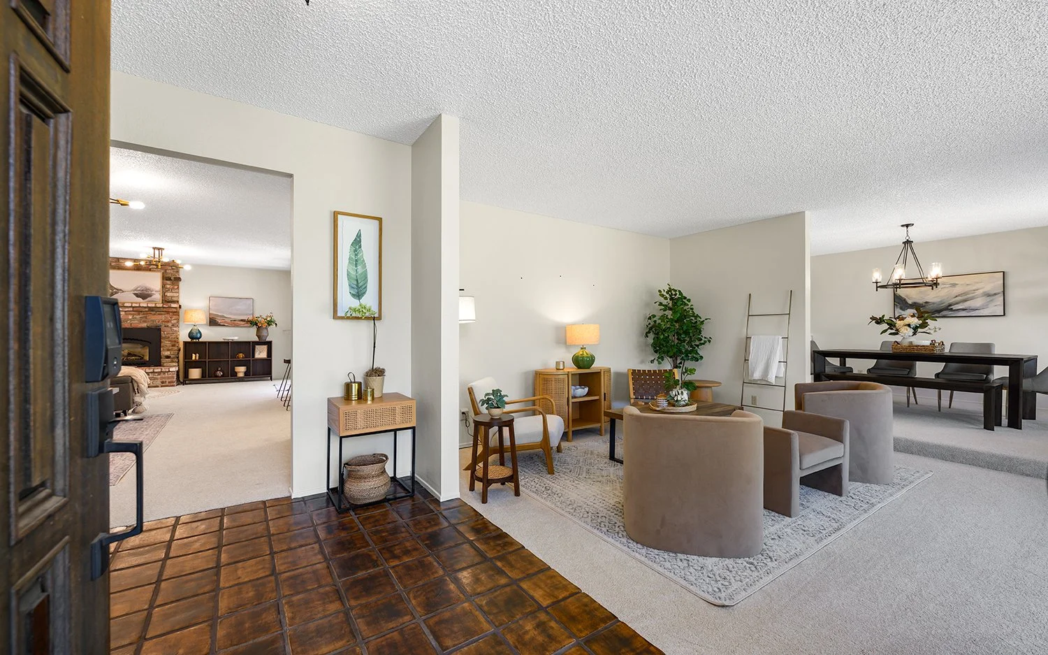 Living room with beige chairs and a small coffee table on a patterned rug, a large plant, a side table with lamps, and a dining area with a table and black chairs in the background.