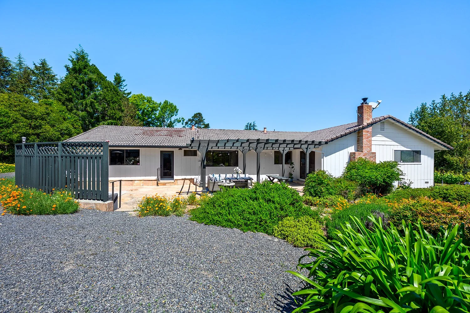 A single-story white house with a dark tiled roof and brick chimney, surrounded by green bushes and plants, with a gravel driveway and a patio area with outdoor furniture under a black pergola, on a sunny day with a clear blue sky.