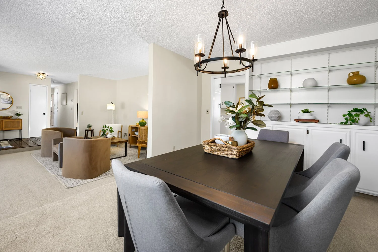 Dining room with a dark wooden table, light gray upholstered chairs, a white built-in bookshelf with decorative vases and plants, and a black metal chandelier with candle-style lights hanging above the table.