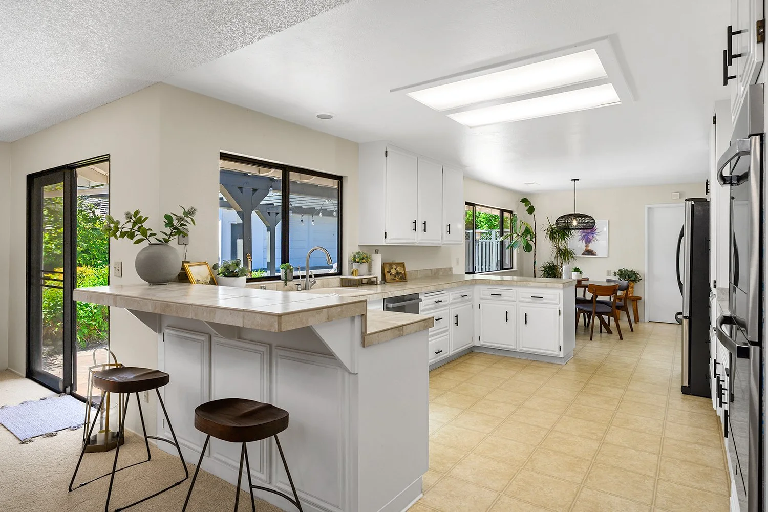 Bright kitchen with white cabinets, beige countertops, large windows, and a small dining area with a plant.