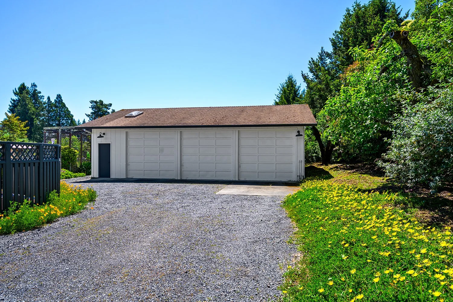 A white garage with a brown roof, two garage doors, a side door, outdoor lights, and surrounded by green bushes and yellow flowers. Gravel driveway leading to the garage under a clear blue sky.