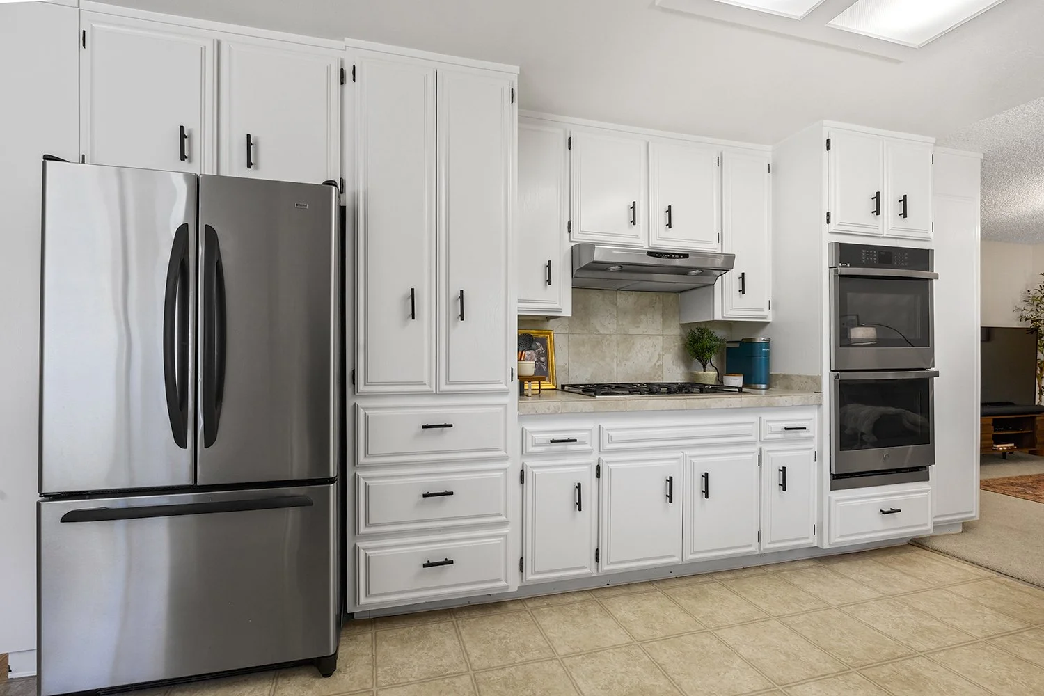 White kitchen cabinets with black handles, stainless steel refrigerator, built-in oven, and stovetop, beige tile backsplash and floor, small potted plant and blue container on the counter.