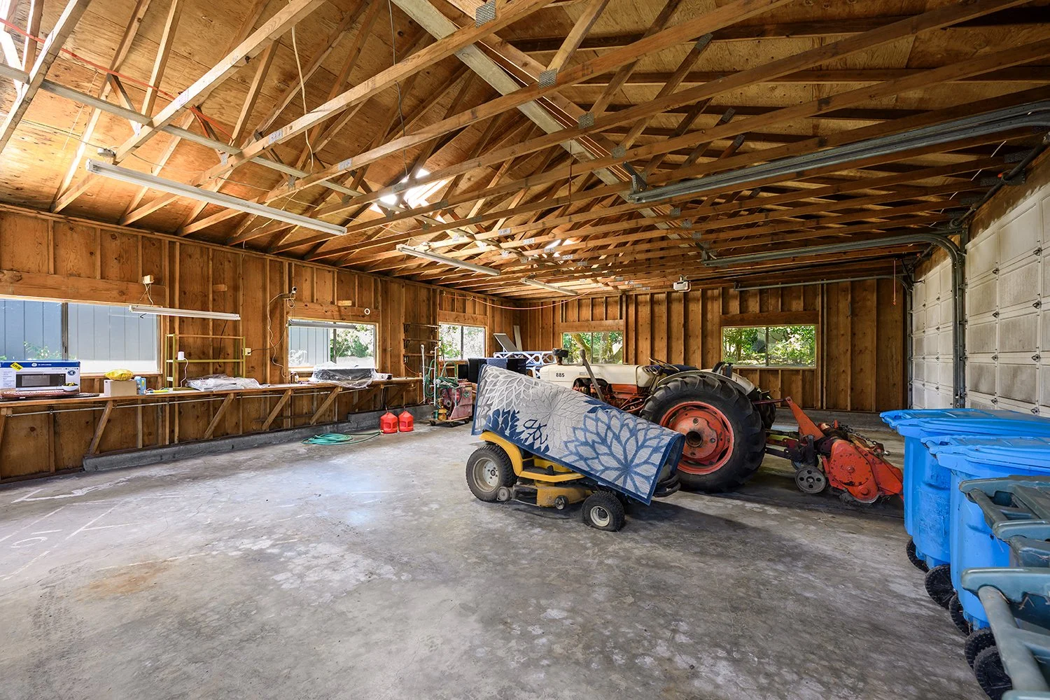 Interior of a wooden garage with a tractor, a ride-on lawn mower, blue storage bins, and a workbench along the wall.