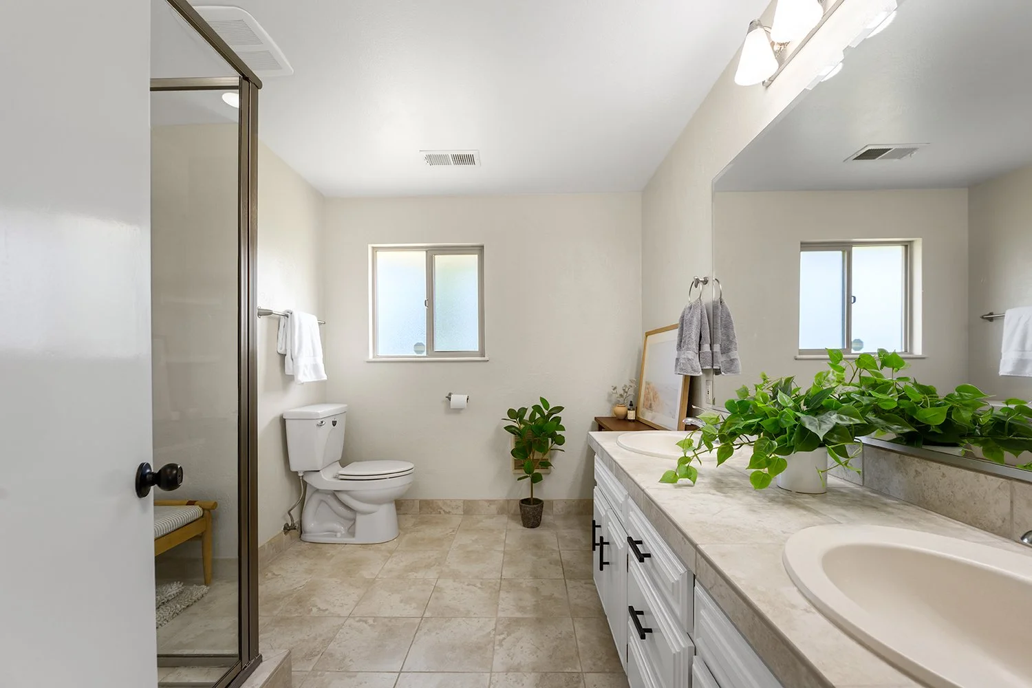 Bright bathroom with white cabinetry, beige countertops, two sinks, large mirror, green plants, window, toilet, towel racks, shower with glass door, small bench, artwork, towels, and decorative items.