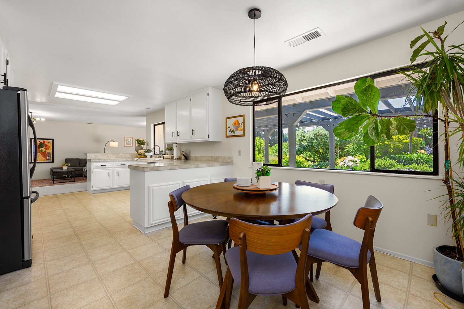 Kitchen and dining area with a round wooden table, six chairs, and large window showing greenery outside.