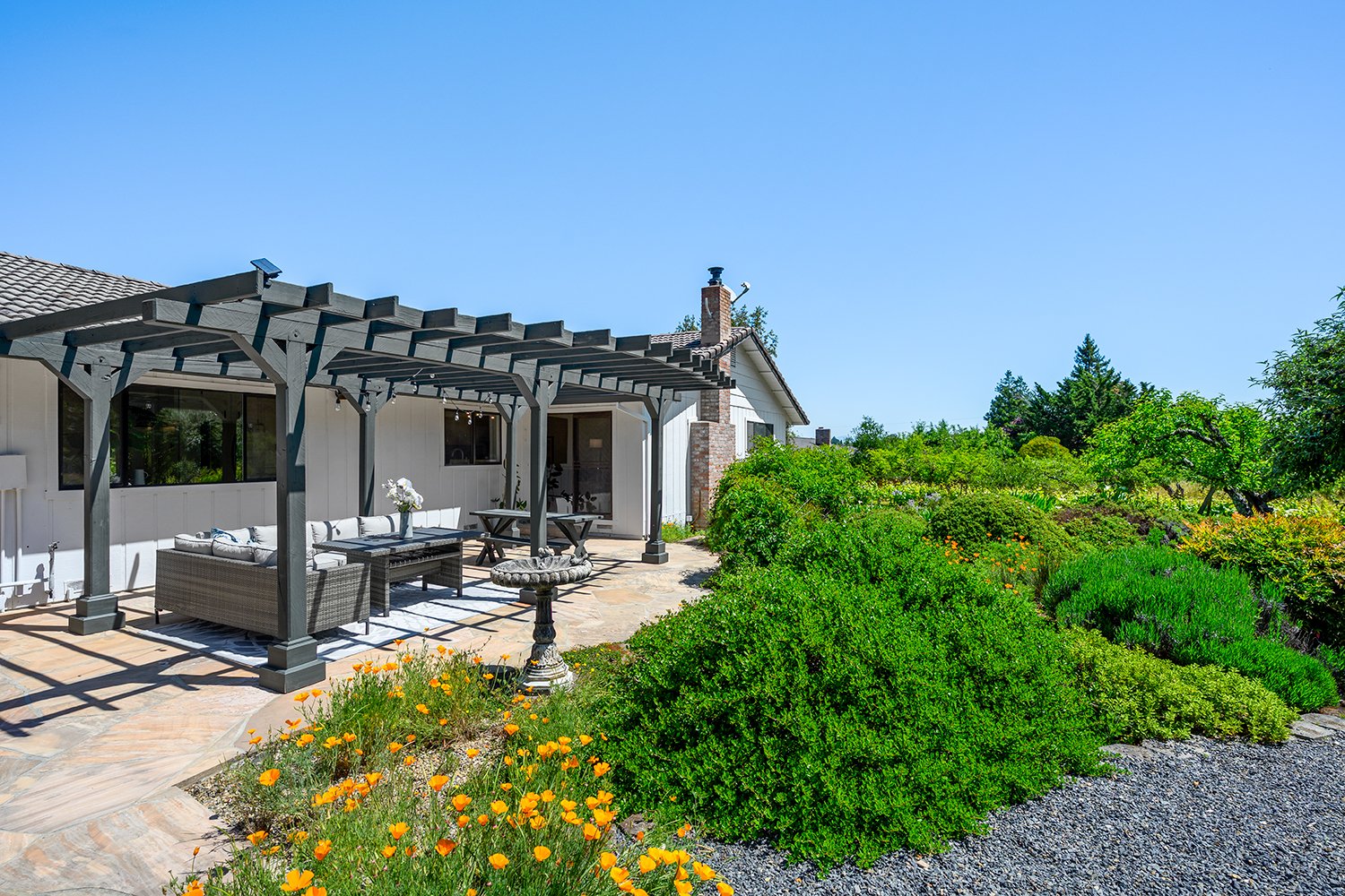 Backyard patio with a pergola, outdoor furniture, and landscaped garden with yellow flowers and green bushes under a clear blue sky.