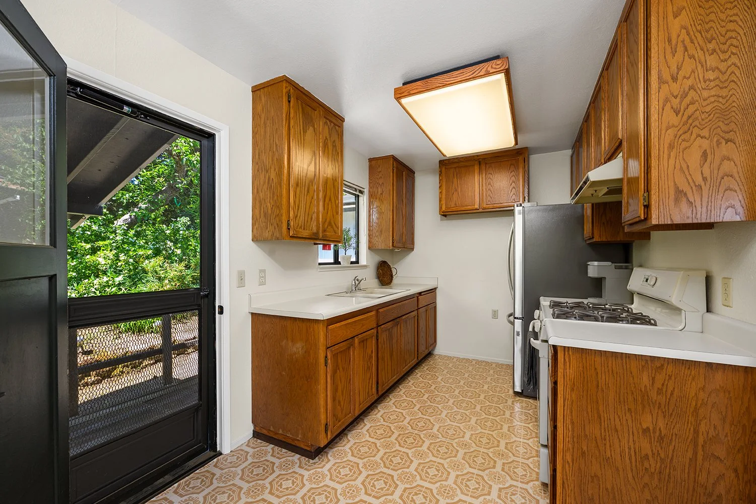 Kitchen with wooden cabinets, white countertop, stove, refrigerator, and a sliding door leading outside to greenery.