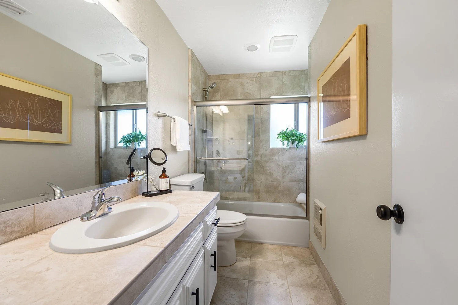 A clean bathroom featuring a beige tiled shower with sliding glass doors, a white toilet, a vanity with a white sink, a large mirror, and two framed abstract artworks on the walls.