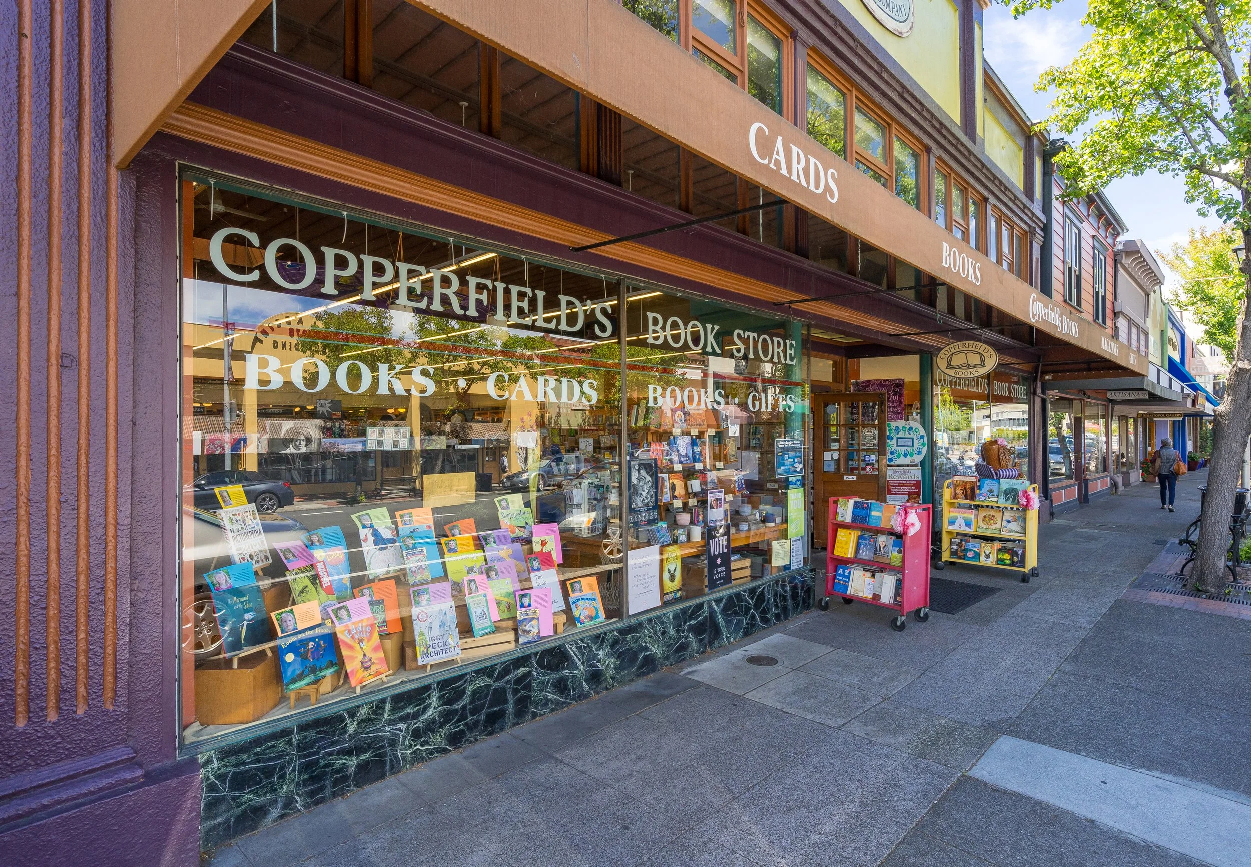 Front view of a bookstore named Copperfield's Books with large glass windows displaying books and greeting cards outside on the sidewalk, a purple wall, and a beige awning that reads 'CARDS' and 'BOOKS'.