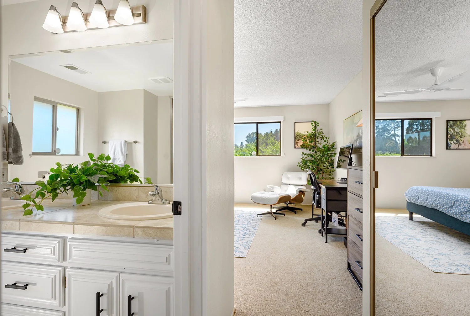 Bathroom with a sink, mirror, and a plant, view into a bedroom with a bed, dresser, chair, and windows showing outdoor trees.