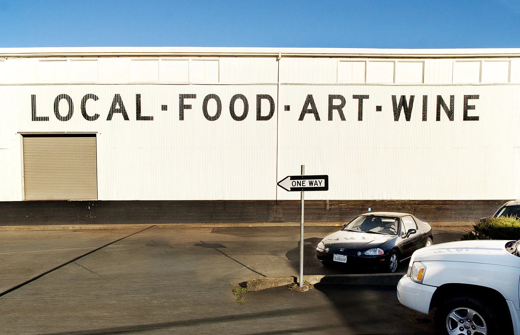 The exterior of a building with large black letters that read 'LOCAL FOOD ART WINE' on a white wall, a small section of a parking lot with cars, including a black and white car, and a one-way street sign pointing to the left against a clear blue sky.