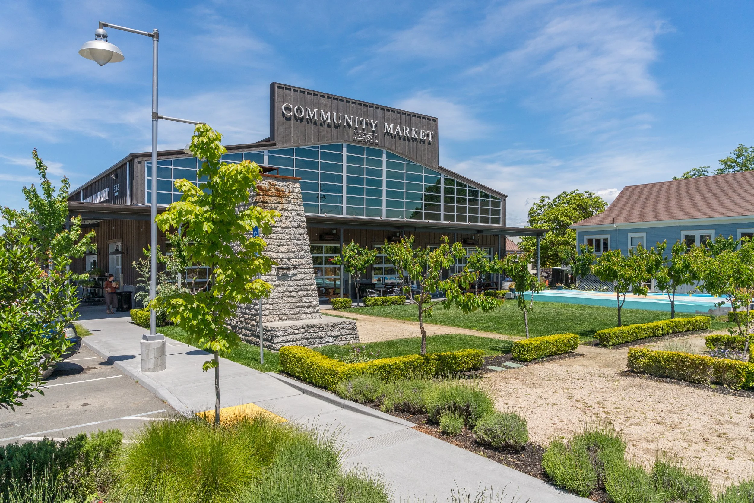 A modern grocery store named 'Community Market' with large windows, surrounded by greenery, under a blue sky with some clouds.
