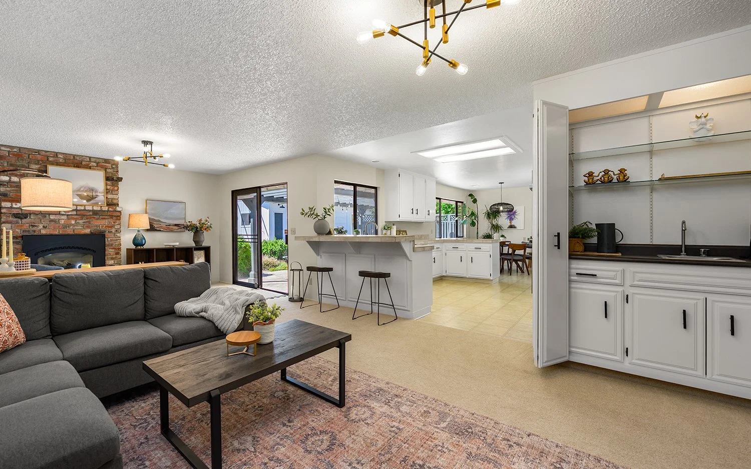 Open-concept living room, kitchen, and dining area with brick fireplace, gray sofa, wooden coffee table, white cabinetry, and sliding glass door to backyard.