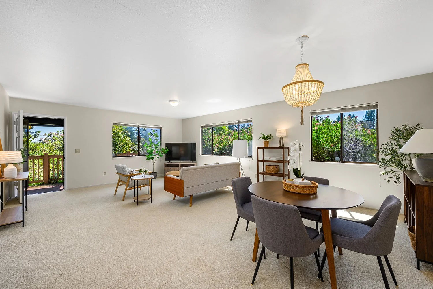 Bright, spacious living room with beige carpet, large windows showing green foliage, a beige sofa, a wooden round dining table with chairs, and various plants and lamps for decor.