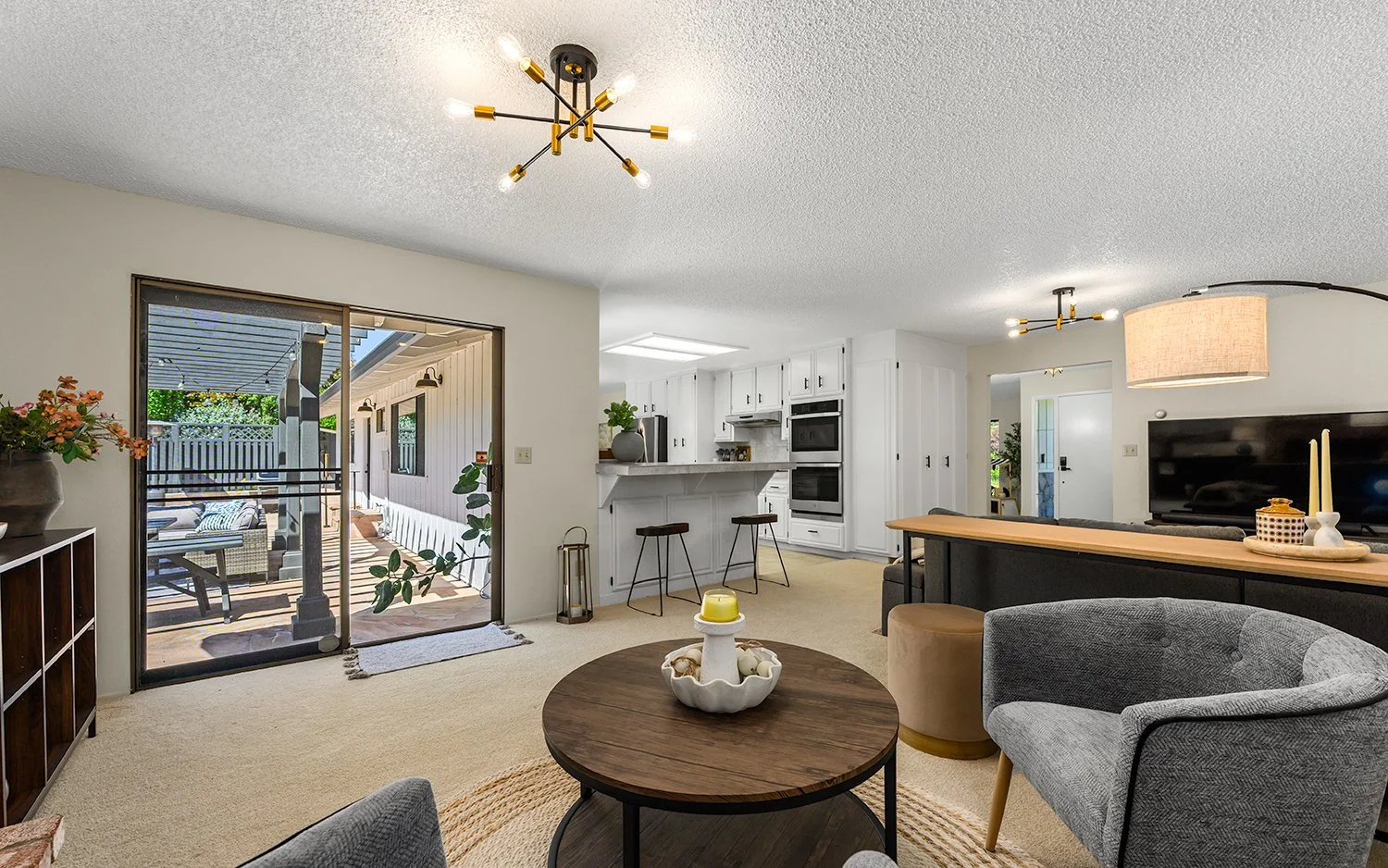 Living room with open sliding door leading to outdoor patio. Interior includes a round coffee table, gray armchair, black and wood TV stand with a large flat-screen TV, and a kitchen with white cabinets, black appliances, and a breakfast bar with thr