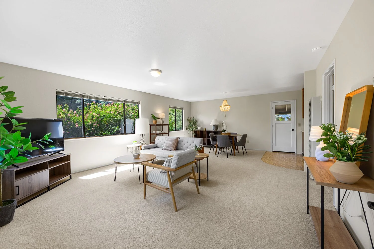 Bright and spacious living room with large windows showcasing greenery outside. Furnished with a television on a wooden stand, a white sofa, an armchair, and a round coffee table. Adjacent to the living space is a dining area with a dark wood table a