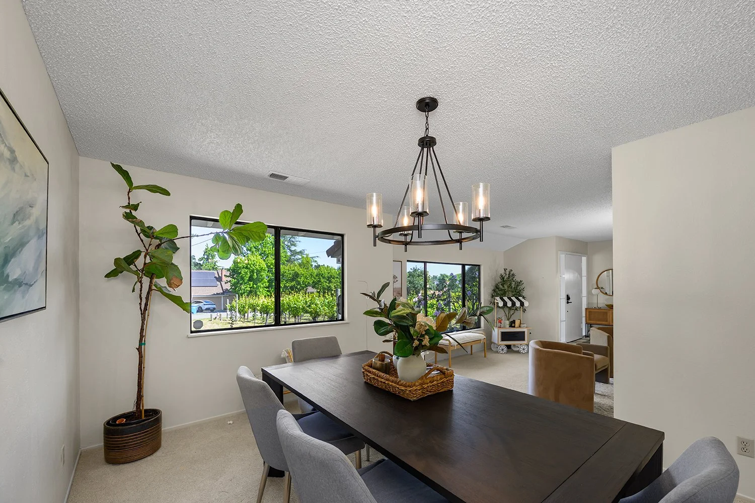 A dining room with a large wooden table and six upholstered chairs. There is a chandelier hanging above the table, a potted plant in a basket on the table, and large windows showing a view of trees and a neighbor's house outside.