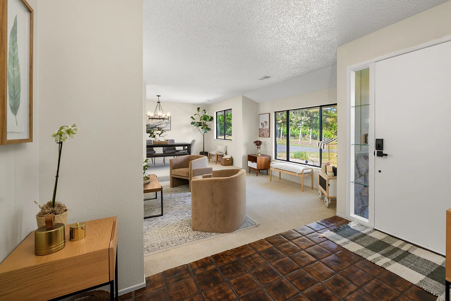 Living room with beige armchairs, a wooden bench, large windows, and artwork on the walls, seen from the entryway with tiled flooring.