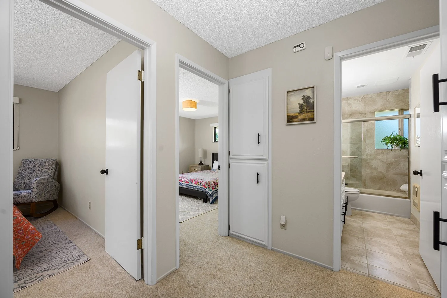 View of hallway with open doors leading to two bedrooms and a bathroom. Bedroom on the left has a chair and a pillow. Bedroom in the middle has a bed with colorful quilt and nightstand. Bathroom on the right has a toilet, a walk-in shower with glass 