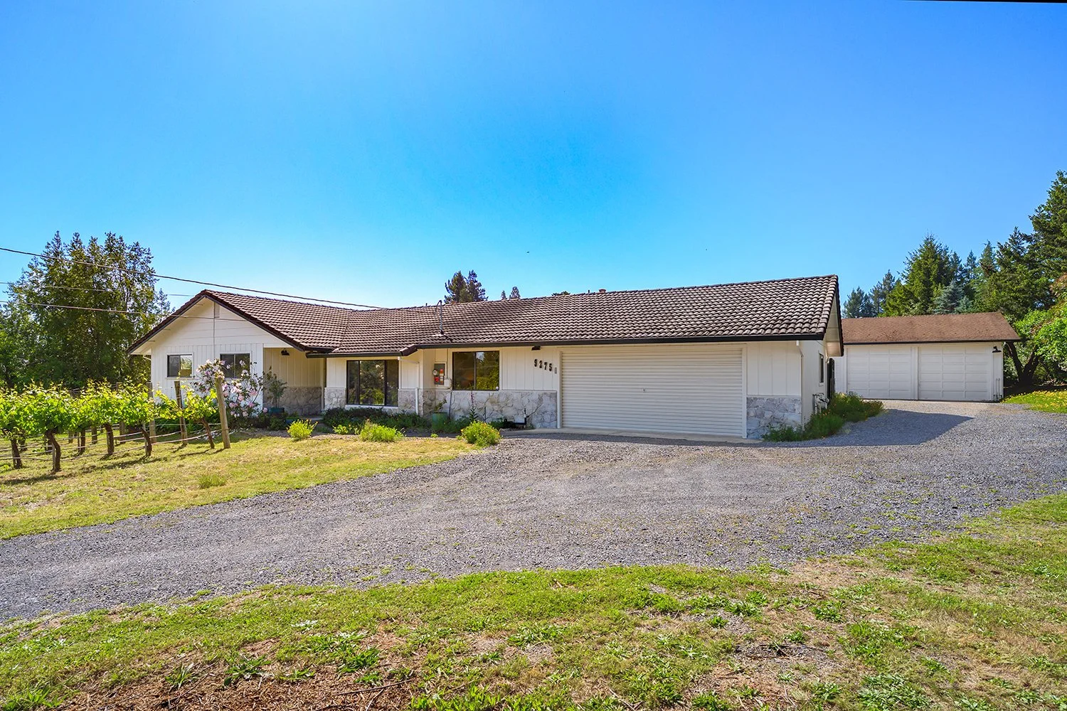 A single-story house with a brown tiled roof, white exterior walls, a large garage door, and a garden with flower bushes and small plants, set on a gravel driveway and green lawn under a clear blue sky.