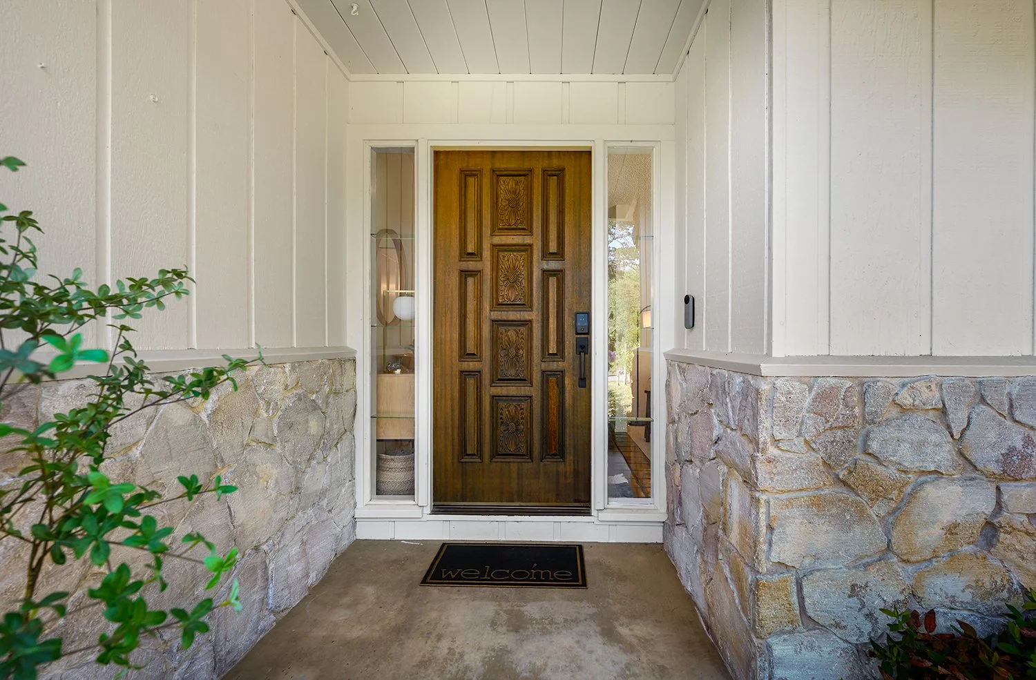 Front porch with a wooden door, stone and siding exterior, welcoming doormat, and greenery on the side.