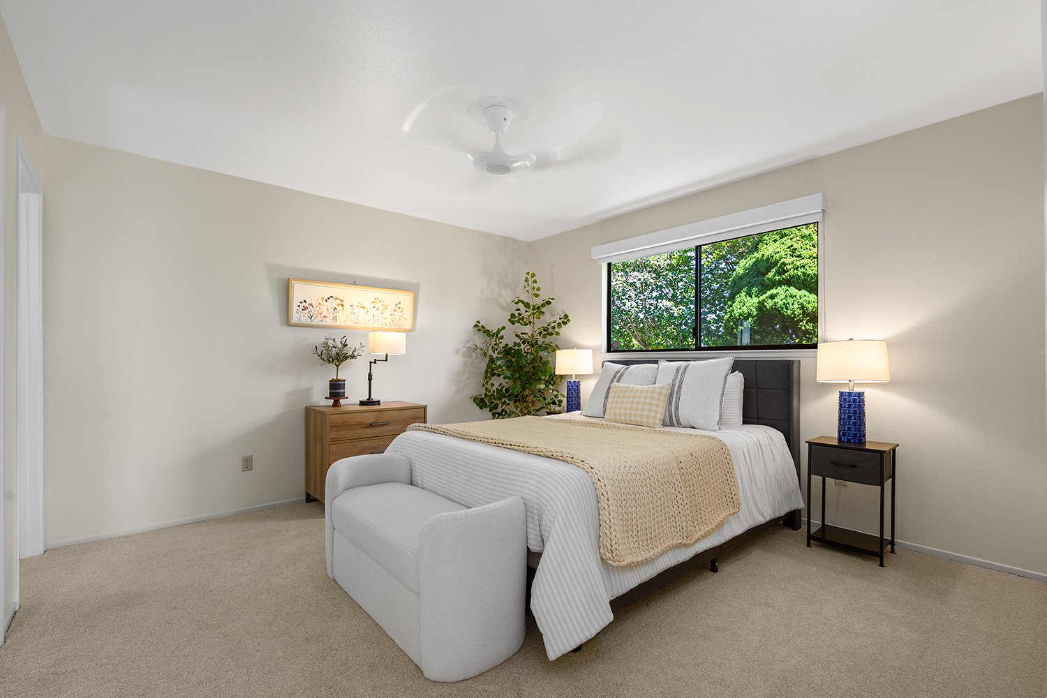 A bedroom with a bed, nightstands with lamps, a window showing green trees outside, a large potted plant, and minimal decorations.
