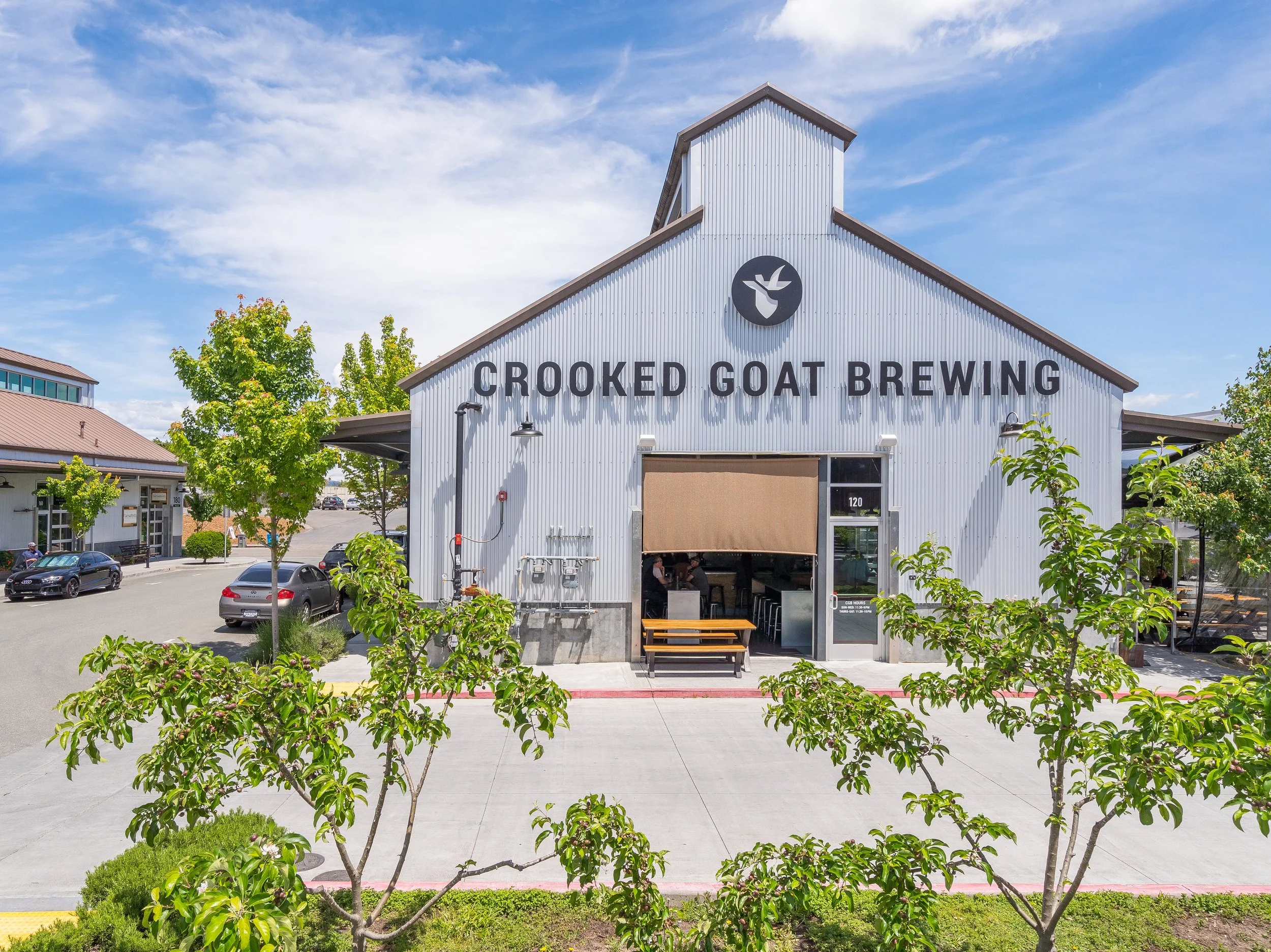 Front view of Crooked Goat Brewing building with a peaked metal roof, surrounded by green trees and a parking lot with cars. The sky is partly cloudy.