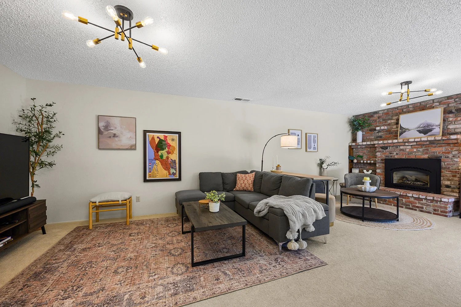 Living room with beige carpeting, a gray sectional sofa with cushions and a throw blanket, a dark wood coffee table with plants, a brick fireplace, and contemporary lighting fixtures.