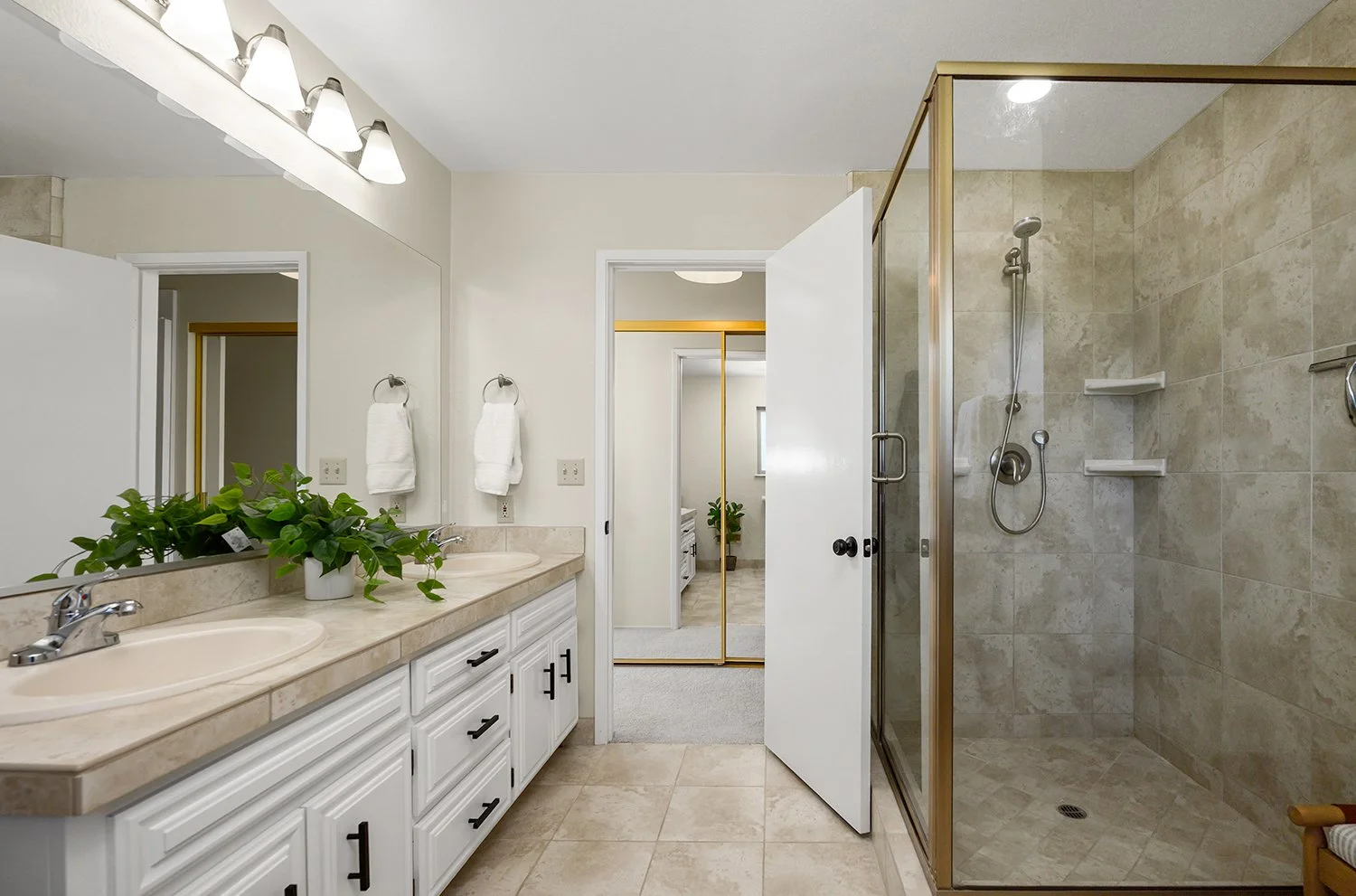 Bathroom with double vanity sinks, marble countertop with plant, large mirror, wall-mounted lights, towels, tiled shower with glass door, shower fixtures, and shelves, beige tiled floor, and open white door leading to adjoining room.