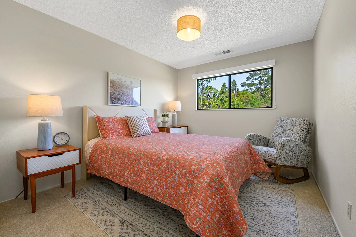A bedroom with a bed covered in an orange patterned quilt, two nightstands with lamps, a patterned armchair, a window with greenery outside, and a framed picture on the wall.