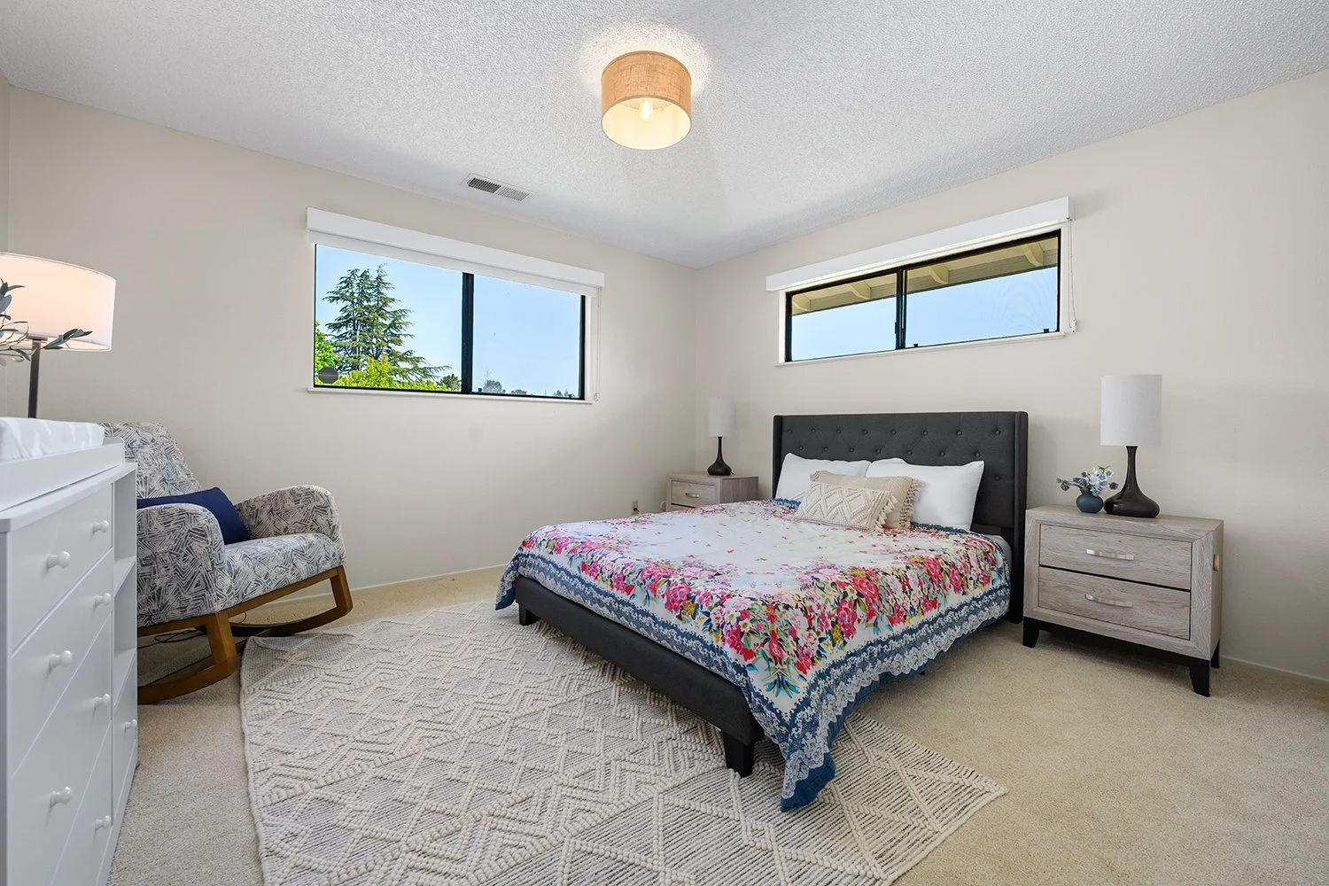 A bedroom with a bed, two nightstands with lamps, a patterned armchair, a dresser, and windows showing greenery outside.