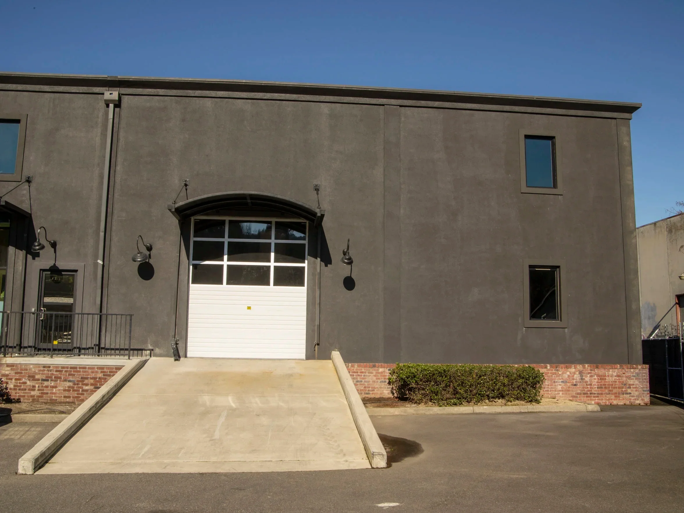 A gray industrial building with garage door, black-framed windows, outdoor wall-mounted lamps, and a concrete slope leading to the garage entrance, set against a clear blue sky with some greenery at ground level.