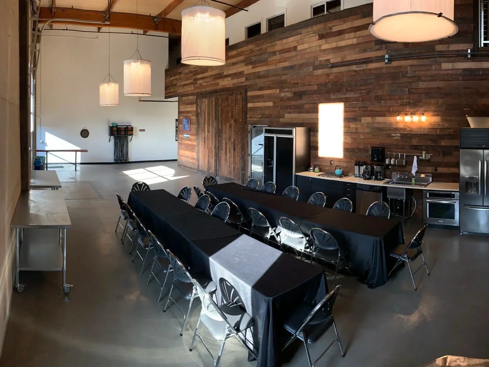 Large gathering space with a long rectangular table covered with a black tablecloth, surrounded by black chairs, set up in a room with a high wooden ceiling, wood-paneled wall, and a kitchenette with stainless steel appliances.