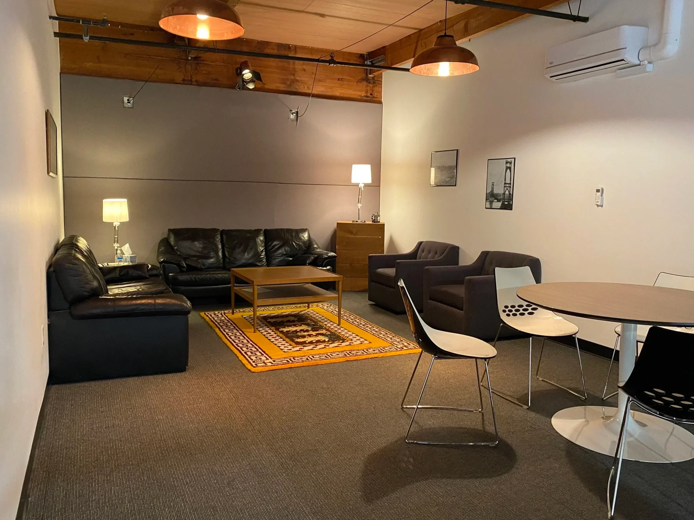 Interior of a living room with black leather sofas, a wooden coffee table, a colorful area rug, and framed pictures on the wall, illuminated by table lamps and pendant lights.