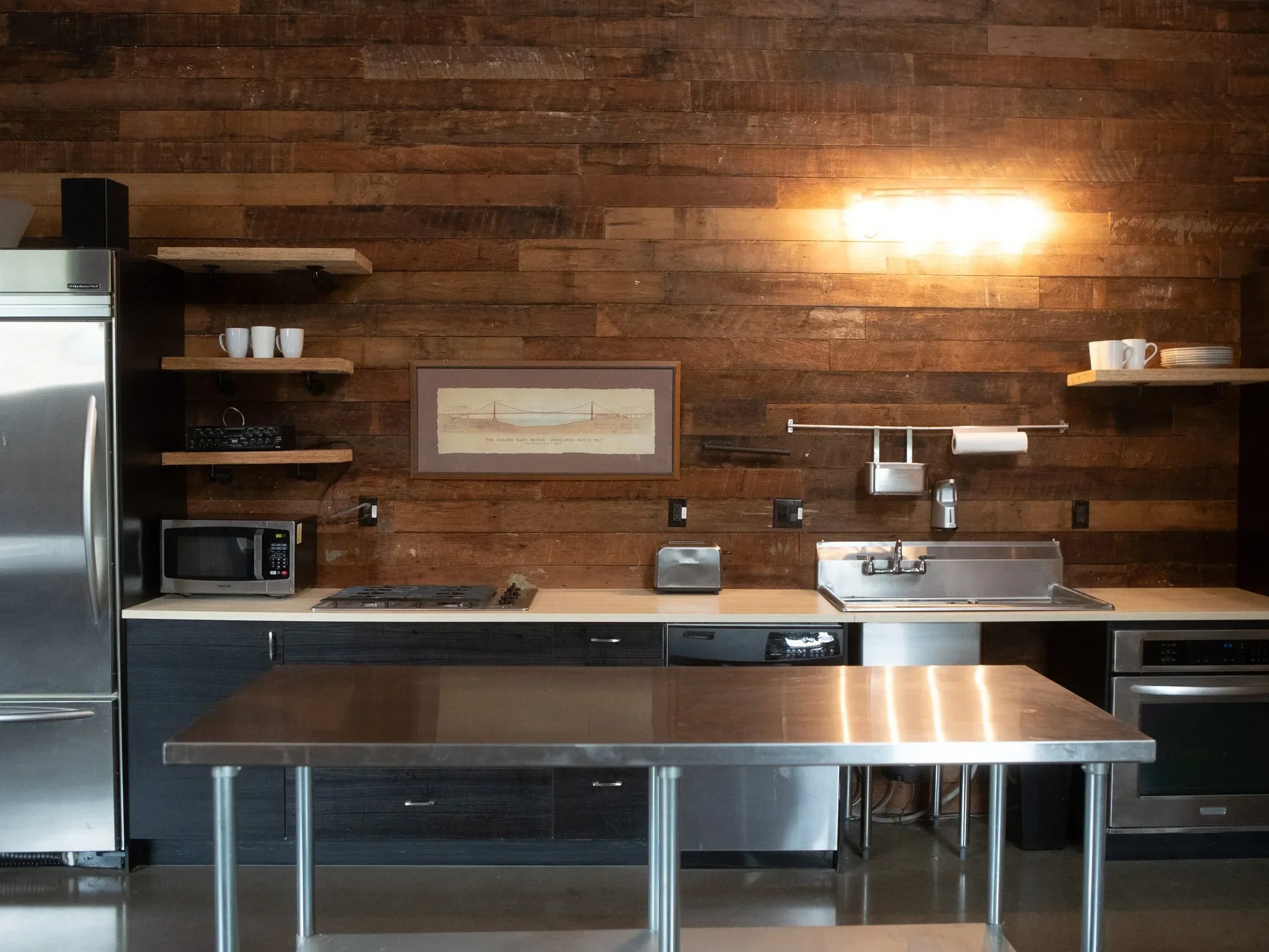 A modern kitchen with a wooden accent wall, stainless steel appliances, open shelving with white mugs and plates, microwave, and a stainless steel sink with a faucet.