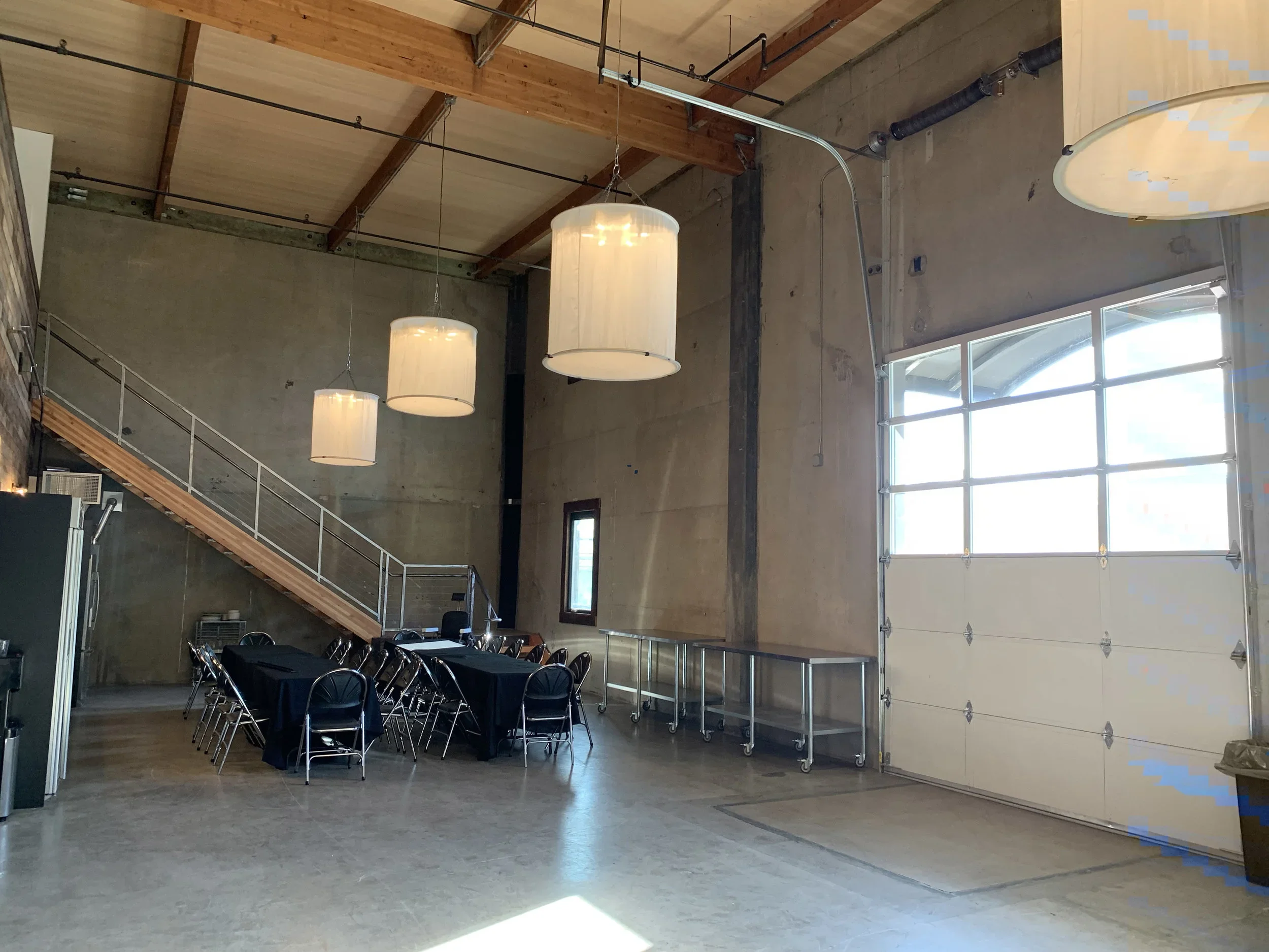 An industrial-style interior with a high ceiling featuring exposed wood beams and black piping. There are three large hanging pendant lights and a row of chairs and tables set up for an event or meeting. A large glass garage door allows natural light